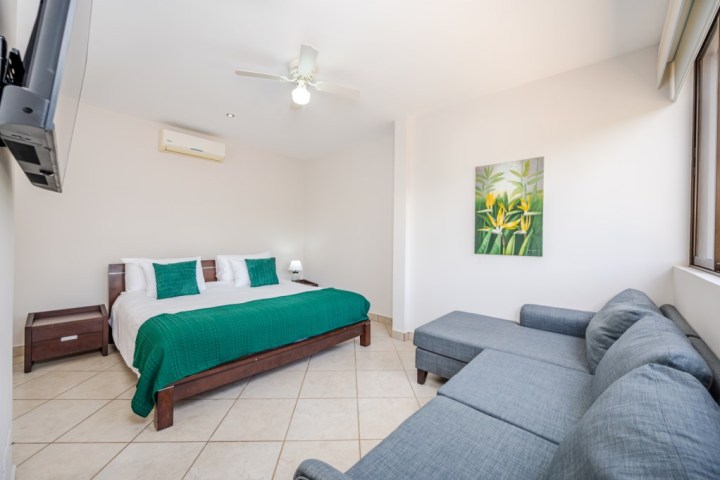 Bedroom with a modern bed, teal accents, gray sofa, ceiling fan, and wall art, featuring neutral tones and tile flooring.