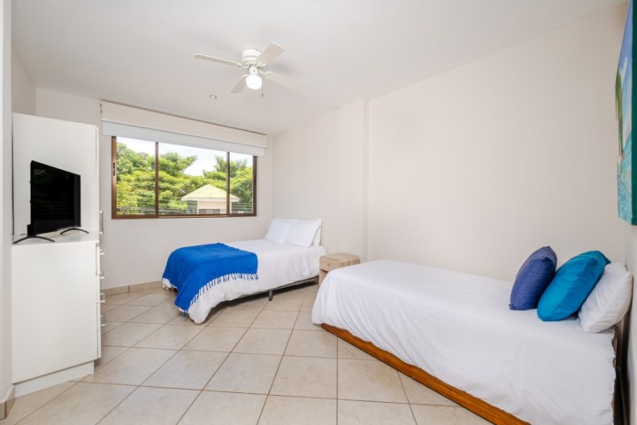 Room with two beds, wall-mounted TV, ceiling fan, and large window with a view of trees.