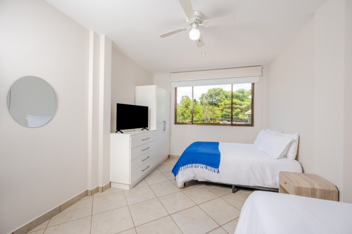 Minimalist bedroom with two beds, blue throw, TV on dresser, ceiling fan, and large window.