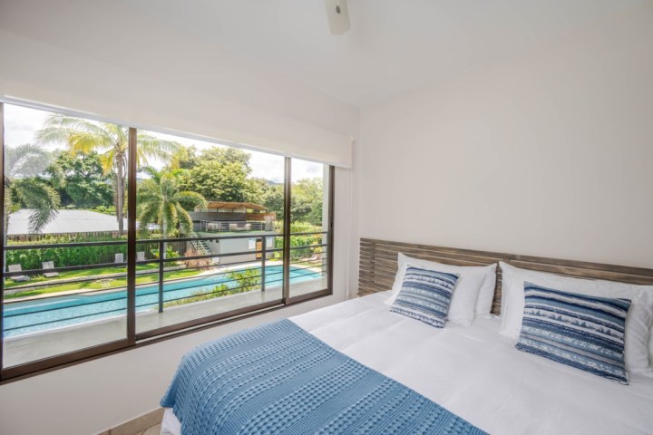 Bedroom with white bedding, blue accents, and a large window view of a pool and palm trees.