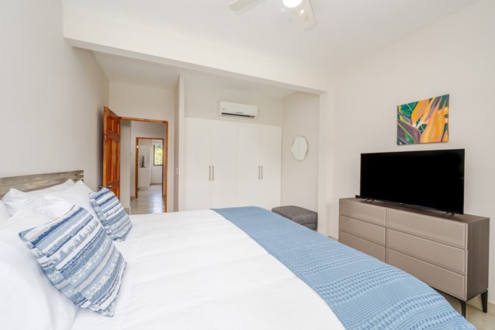 Modern bedroom with a bed, blue accents, dresser, TV, artwork, and a door leading to a hallway.