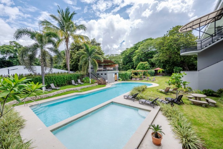 Outdoor pool with lounge chairs, palm trees, and a garden under a partly cloudy sky.