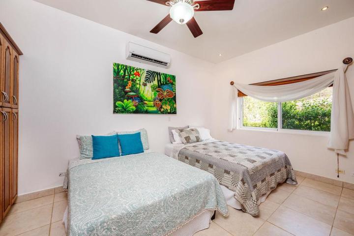 Bedroom with two beds, colorful artwork, wooden cabinet, ceiling fan, and a large window with a garden view.
