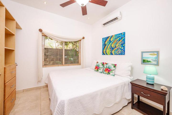 Small bedroom with a white bed, colorful pillows, and artwork, wooden shelves, and a lamp on a bedside table.