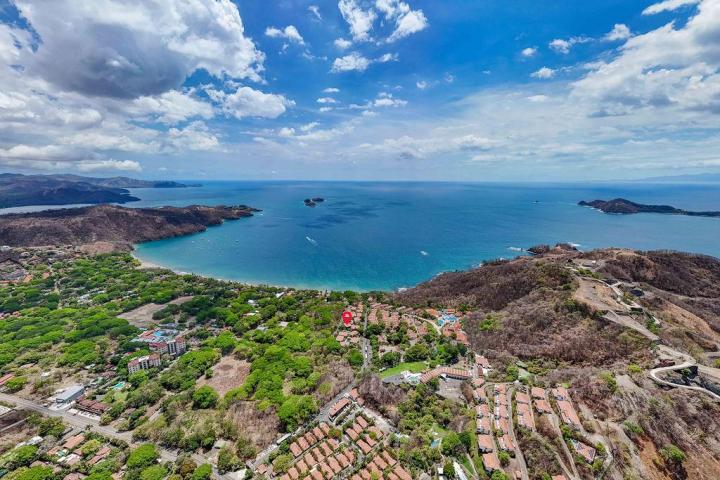 Aerial view of a coastal town with sandy beach, greenery, and blue ocean under a partly cloudy sky.