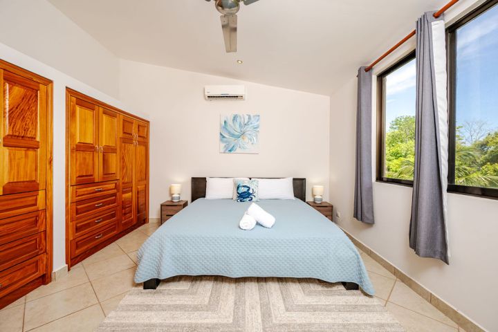 Bedroom with blue bedding, wooden wardrobes, large window, and two bedside tables with lamps.