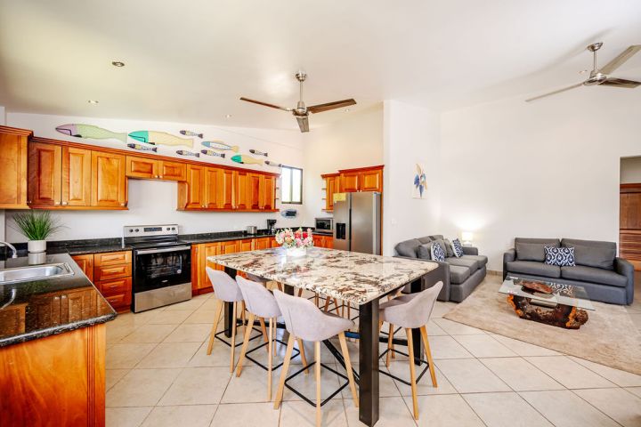 Modern kitchen and living area with wooden cabinets, island, sofas, and ceiling fans.