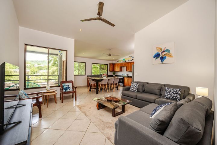Modern living room with gray sofas, TV, dining area, and kitchen. Large windows offer views of greenery.