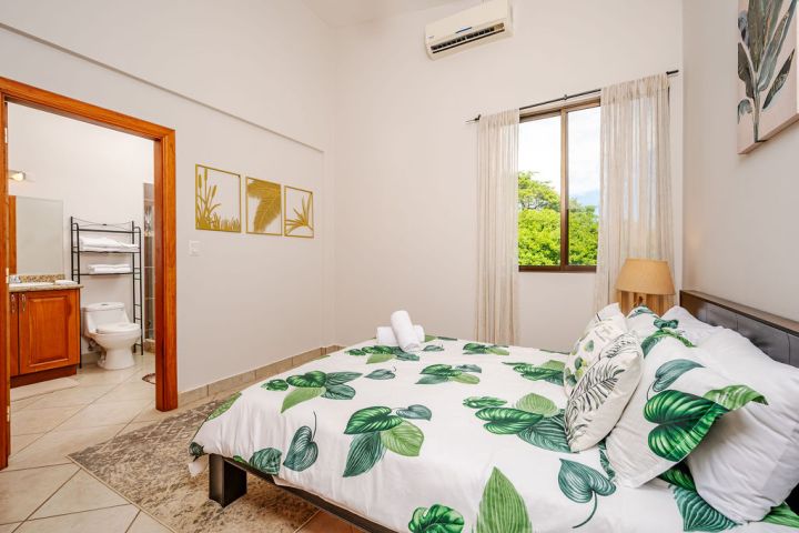 Bedroom with leaf-patterned bedding, window, lamp, and open door to bathroom, featuring plants and neutral decor.