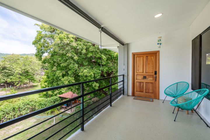 Covered balcony with two turquoise chairs, wooden door, and view of trees.