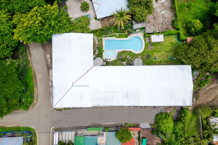 Aerial view of a large white-roofed building, pool, and surrounding greenery.