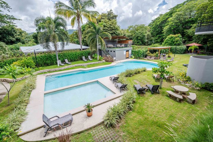 Outdoor pool area with deck chairs, palm trees, and greenery on a cloudy day.