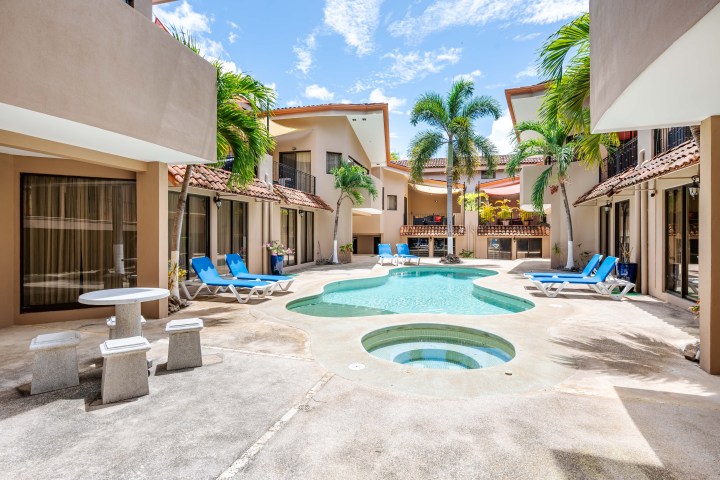 Outdoor pool area with lounge chairs and palm trees surrounded by two-story buildings.