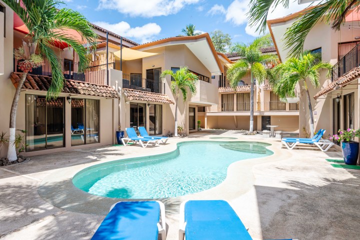 Courtyard with pool, palm trees, and sun loungers between tan buildings under a blue sky.