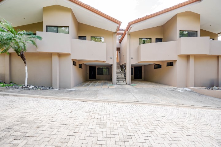 Two beige modern houses with shared staircase, palm tree, and paved driveway.