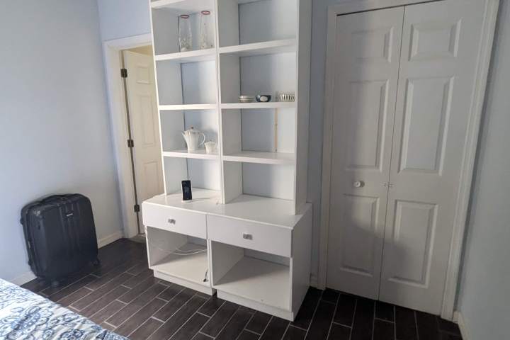 Room with white shelving, a bed with blue floral sheets, and a black suitcase on the floor.