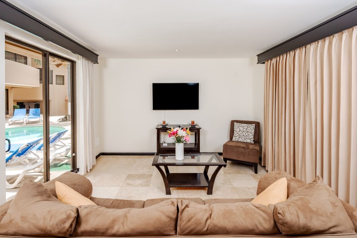 Modern living room with brown couch, TV, glass table, and view of pool through large windows.