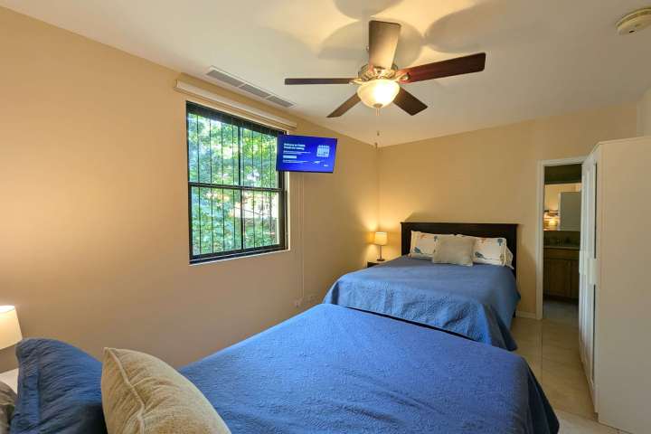 Bedroom with two blue-covered beds, ceiling fan, TV, and a window showing outdoor greenery.