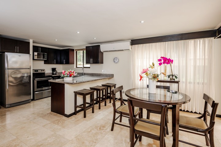 Modern kitchen with dark cabinets, island, and dining table with chairs. Bright with flowers and large window.