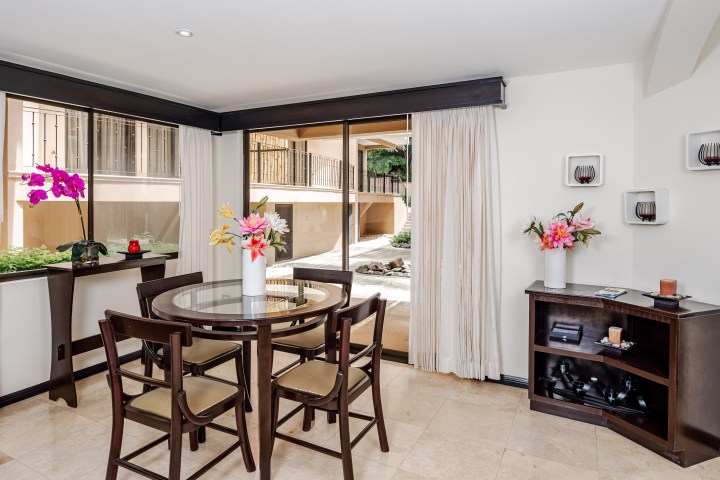 Dining area with dark wood table, chairs, and floral centerpieces, near large windows and shelves.