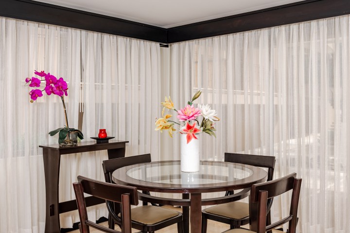 Dining room with glass table, wooden chairs, vase of flowers, and sheer curtains.