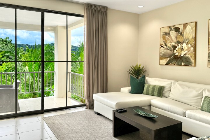 Living room with white sofa, green cushions, art, and balcony view of trees and hills.