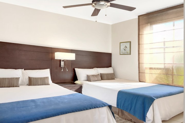 Two beds with blue throws, modern room with ceiling fan and window.