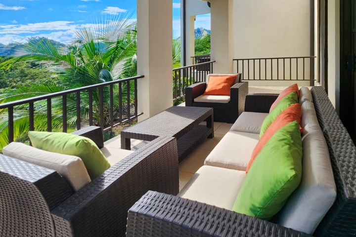 Outdoor patio with wicker furniture, green and orange cushions, overlooking lush greenery.