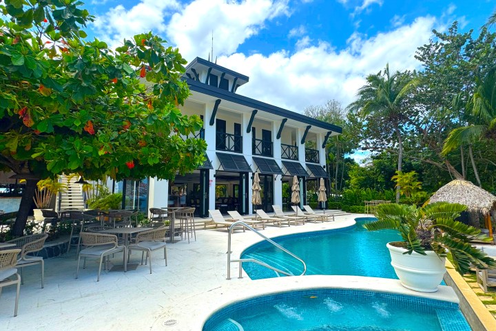 Two-story building by a pool with chairs, tables, and lush greenery under a blue sky.