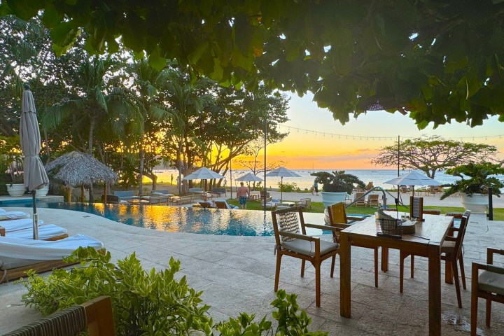 Outdoor dining area by pool with sunset ocean view, surrounded by trees and greenery.