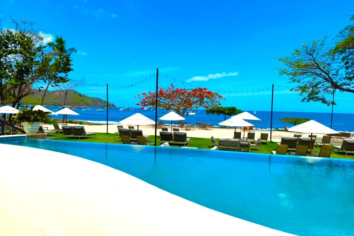 Infinity pool overlooking a beach with lounge chairs and white umbrellas under a clear blue sky.