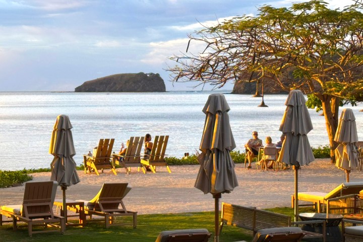 Beachside chairs and umbrellas overlooking ocean, with distant island and tree in the foreground.