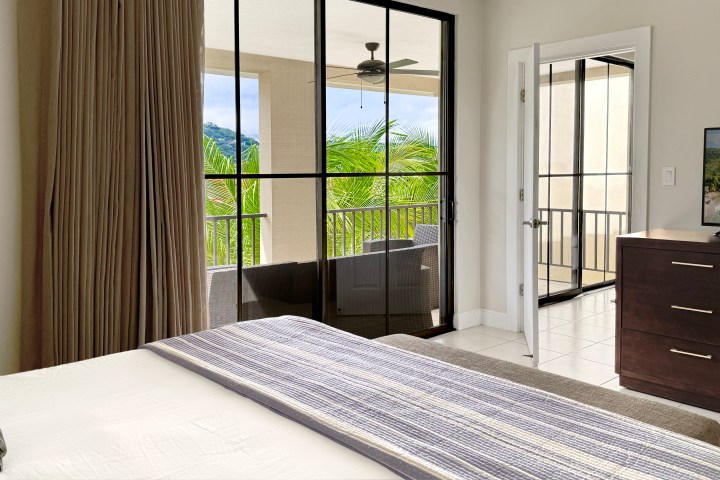Bedroom with bed, dresser, TV, and open balcony door showing plants and railing.