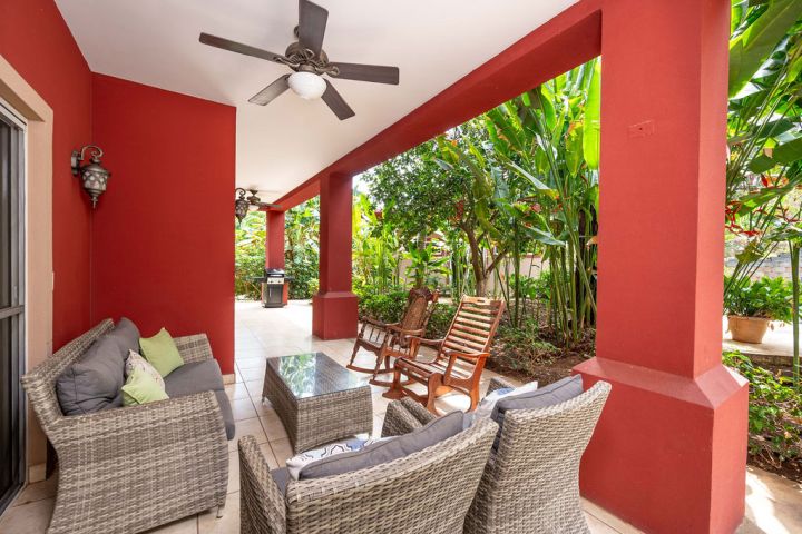 Covered red patio with ceiling fan, wicker furniture, glass table, and garden view.