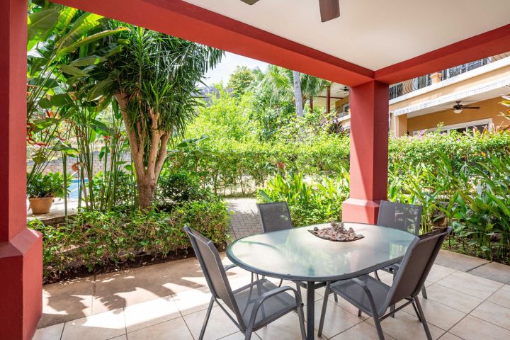 Outdoor patio with red pillars, glass table, four chairs, surrounded by lush green plants.