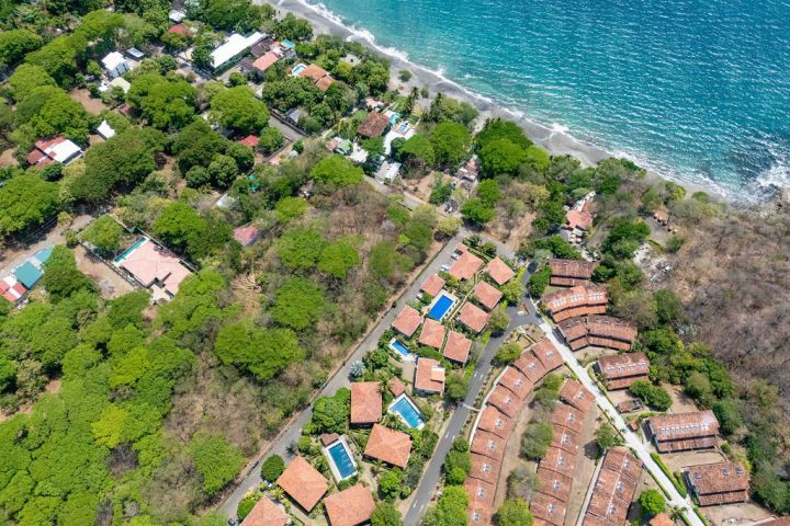 Aerial view of coastal resort with swimming pools, villas, and lush trees near the ocean.
