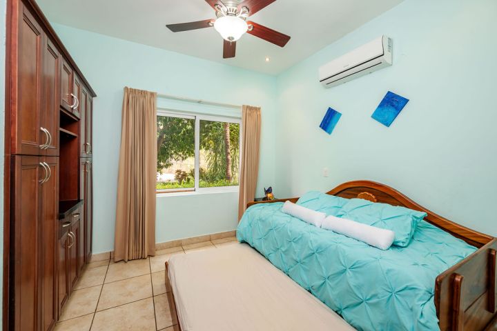 Bedroom with a teal bed next to a window, wooden cabinet, ceiling fan, and two blue wall paintings.