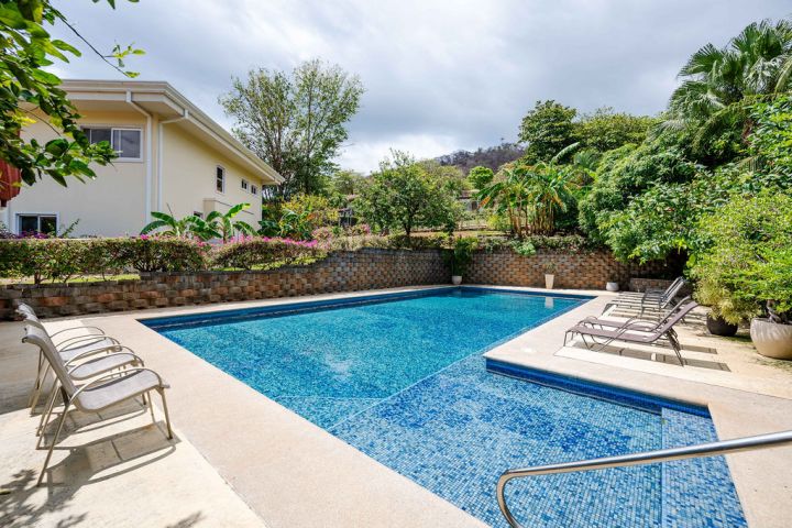Outdoor swimming pool with lounge chairs, surrounded by greenery and a house in the background.
