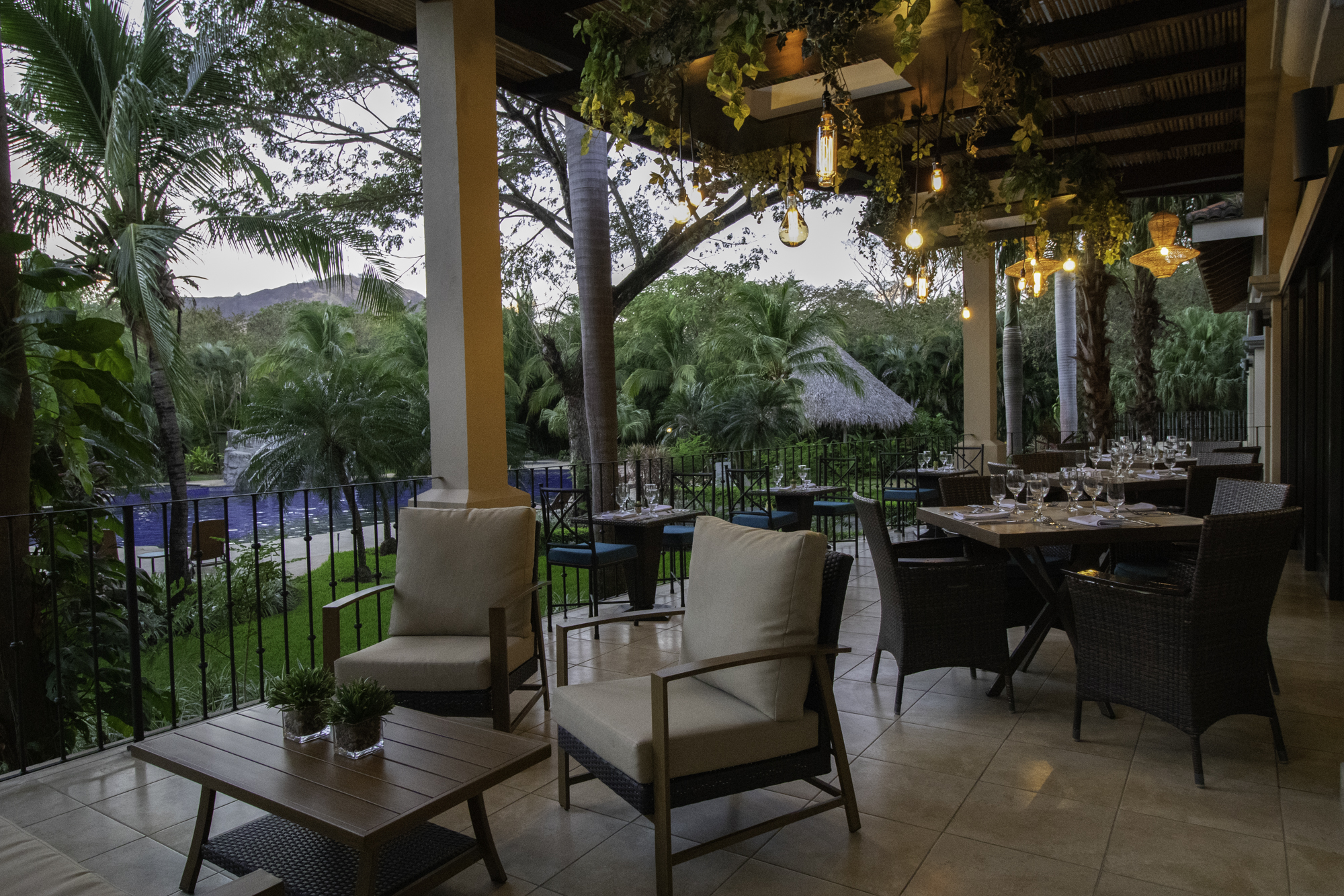 Sugoi, Playas del Coco Outdoor patio with seating, dining tables, and hanging lights overlooking lush greenery and a pool.