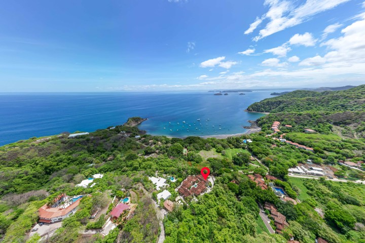 Aerial view of lush coastline with blue ocean, boats, and scattered buildings below a clear sky.