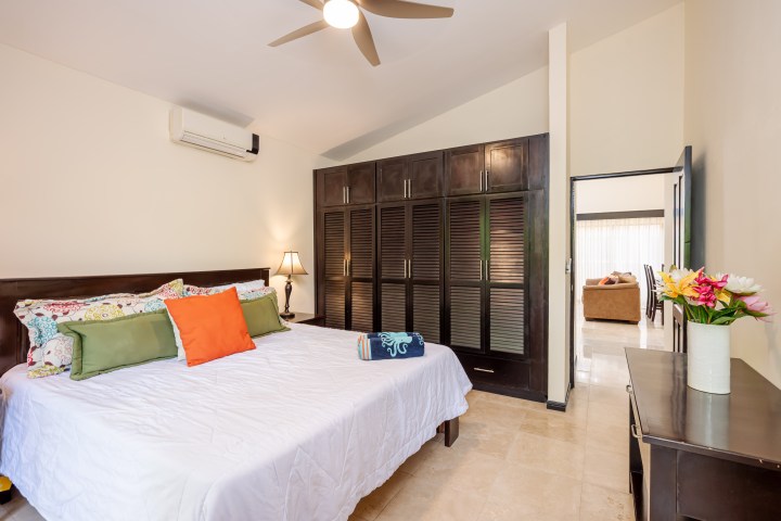 Bedroom with bed, colorful pillows, ceiling fan, wardrobe, and vase of flowers on a dresser.