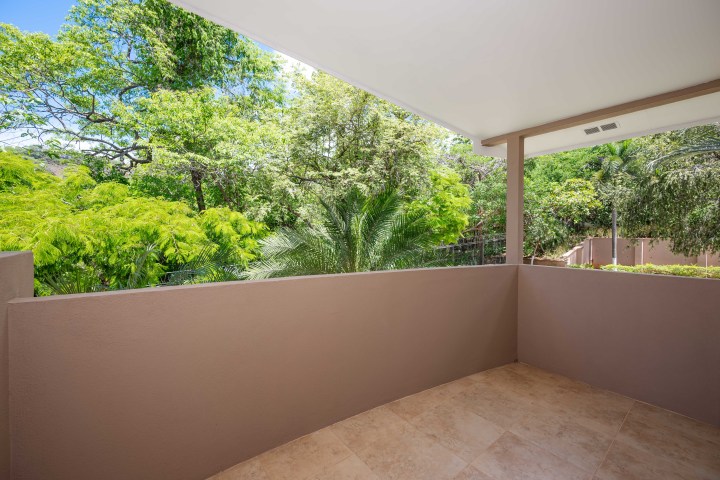 Covered balcony with tan walls, tile floor, and view of lush green trees.