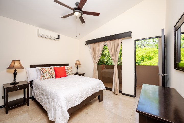 Bedroom with a white bedspread, ceiling fan, and open doors to a balcony with trees visible outside.