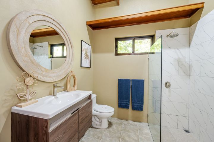 Bathroom with round mirror, sink, toilet, shower, and two blue towels on the wall.