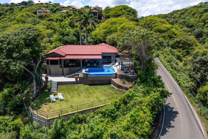 Hilltop house with red roof, pool, green lawn, surrounded by lush trees and adjacent to a winding road.