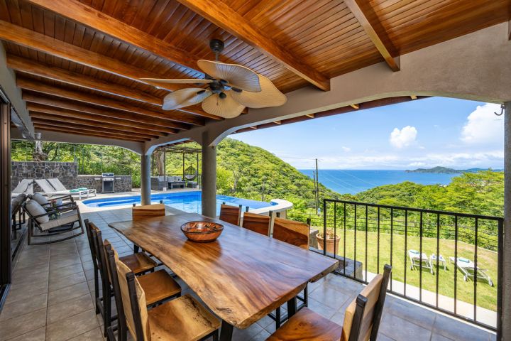 Covered patio with wooden table, ceiling fan, pool, and ocean view.