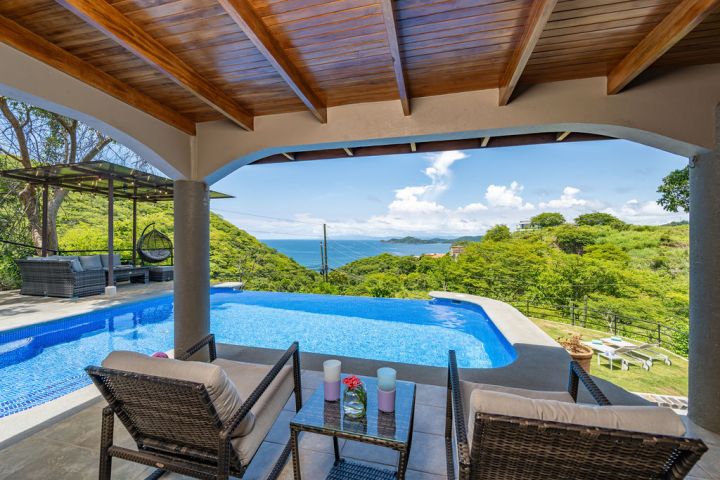Covered patio with wicker chairs, infinity pool, ocean and hillside view.