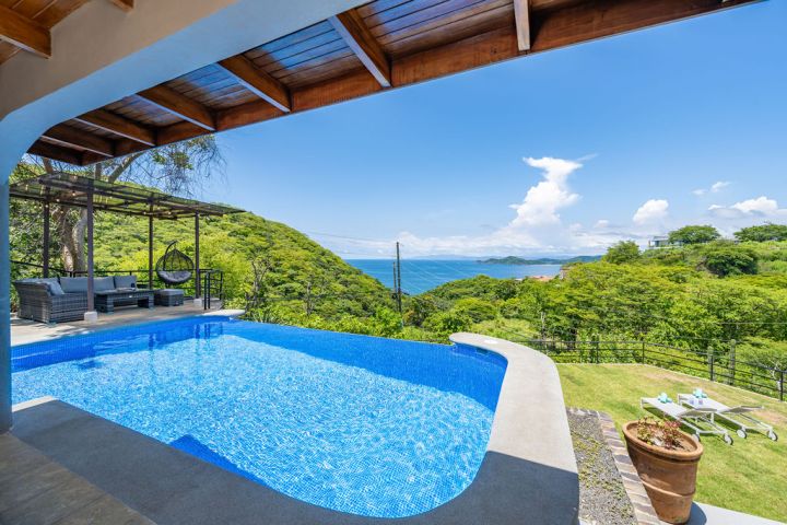 Covered poolside patio overlooking lush hills and ocean under a clear blue sky.