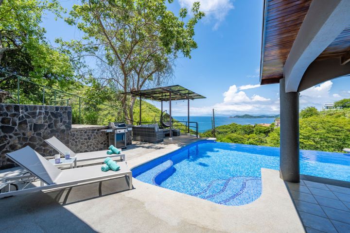 Infinity pool with lounge chairs and ocean view under a clear sky.