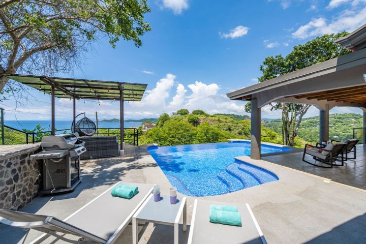 Outdoor pool area with loungers, grill, and ocean view under a blue sky.